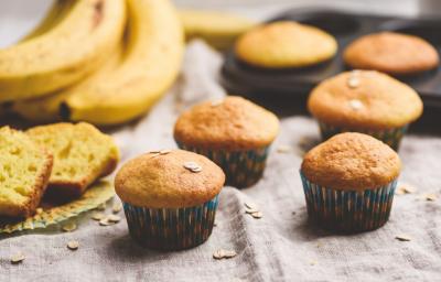Fotografia de quatro muffins de banana e aveia dentro de uma forminha de papel colorida sobre uma toalha de mesa em tons de bege. Ao lado, um cacho de bananas, duas fatias do bolo, e ao fundo, dois muffins em uma forma preta de bolinhos. Fotografia de quatro muffins de banana e aveia dentro de uma forminha de papel colorida sobre uma toalha de mesa em tons de bege. Ao lado, um cacho de bananas, duas fatias do bolo, e ao fundo, dois muffins em uma forma preta de bolinhos.