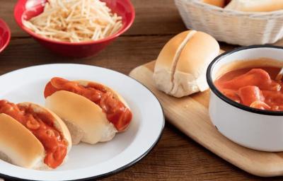 Fotografia em tons de marrom e vermelho de uma bancada de madeira com um prato branco, sobre ele dois cachorros quentes. Ao lado uma tábua de madeira sobre ela uma panela branca com salsicha ao molho e pães. Ao fundo dois recipientes vermelhos.