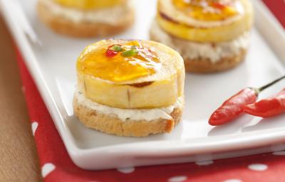 Fotografia em tons de vermelho em uma bancada de madeira, uma toalha vermelha com bolinhas brancas, um recipiente branco retangular com os canapés de banana com geleia de pimenta. Fotografia em tons de vermelho em uma bancada de madeira, uma toalha vermelha com bolinhas brancas, um recipiente branco retangular com os canapés de banana com geleia de pimenta.
