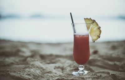 Fotografia de uma taça com um coquetel de água com gás e frutas, e na borda, metade de uma fatia de abacaxi. Dentro da bebida tem um canudo preto, na qual está apoiada sobre a areia.