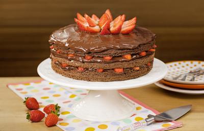 Fotografia em tons de marrom e vermelho de uma bancada de madeira, uma boleira com o bolo de chocolate e morangos. Ao lado quatro morangos para decorar, um espátula para servir e três pratos coloridos em tons de laranja com garfos. Fotografia em tons de marrom e vermelho de uma bancada de madeira, uma boleira com o bolo de chocolate e morangos. Ao lado quatro morangos para decorar, um espátula para servir e três pratos coloridos em tons de laranja com garfos.