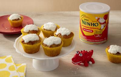 Fotografia em tons de amarelo e vermelho com 5 bolinhos ao centro. Por cima de cada bolinho existe um creme de leite de coco, coco ralado e NINHO Primeira Infância