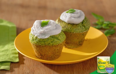 Muffins de espinafre sobre prato amarelo, com cobertura cremosa clara e folhas verdes por cima, dispostos em mesa de madeira.