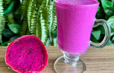 Fotografia mostra um copo com bebida rosa, ao lado metade de uma pitaya aberta.