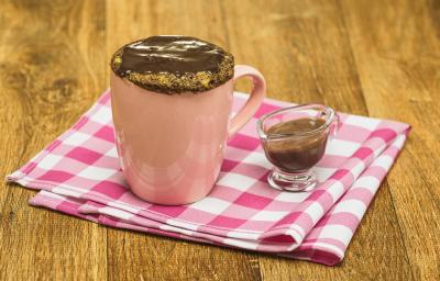 Fotografia em tons de rosa em uma bancada de madeira escura, um pano rosa xadrez, uma caneca rosa com um bolo de caneca de banana com mel, aveia e cobertura de chocolate. Ao lado, um potinho de vidro com a calda de chocolate.