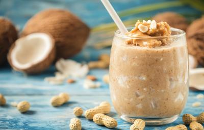 Fotografia de um copo de vidro com uma receita de batida com coco e paçoca com gelo e amendoim. O copo está sobre uma mesa de madeira azul com alguns amendoins com casca espalhados, e ao fundo, tem alguns cocos inteiros e outras metades. Fotografia de um copo de vidro com uma receita de batida com coco e paçoca com gelo e amendoim. O copo está sobre uma mesa de madeira azul com alguns amendoins com casca espalhados, e ao fundo, tem alguns cocos inteiros e outras metades.