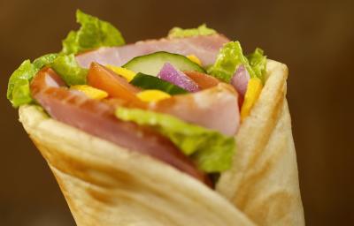 Fotografia de um lanche fechado com pão sírio, alface, tomate, pepino, cebola, molho e peito de peru, e um fundo escuro. Fotografia de um lanche fechado com pão sírio, alface, tomate, pepino, cebola, molho e peito de peru, e um fundo escuro.