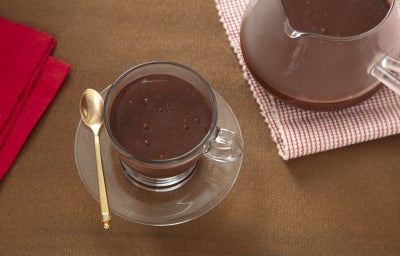 Fotografia de uma xícara de vidro sobre um prato pequeno, também de vidro. Dentro da xícara tem chocolate quente cremoso de nescau extra cacau.