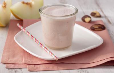Fotografia em tons de branco e rosa de uma bancada branca com um paninho rosa e um prato quadrado branco, sobre ele um copo de vidro com o Smoothie e um canudo listrado rosa. Ao fundo duas fatias de melão e castanhas-do-pará. Fotografia em tons de branco e rosa de uma bancada branca com um paninho rosa e um prato quadrado branco, sobre ele um copo de vidro com o Smoothie e um canudo listrado rosa. Ao fundo duas fatias de melão e castanhas-do-pará.