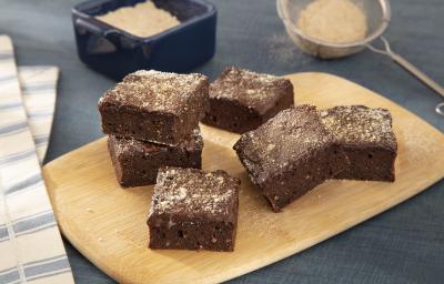 Foto da receita de Brownie com Farinha Láctea NESTLÉ. Observa-se uma tabua de madeira com 6 brownies quadrados com farinha láctea polvilhada Foto da receita de Brownie com Farinha Láctea NESTLÉ. Observa-se uma tabua de madeira com 6 brownies quadrados com farinha láctea polvilhada