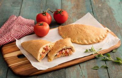 Fotografia em tons de vermelho em uma bancada de madeira com um pano vermelho com bolinhas brancas, uma tábua de madeira, um pape, manteiga em cima e calzones de queijo apoiados sobre ele. Ao fundo, tomates pequenos. Fotografia em tons de vermelho em uma bancada de madeira com um pano vermelho com bolinhas brancas, uma tábua de madeira, um pape, manteiga em cima e calzones de queijo apoiados sobre ele. Ao fundo, tomates pequenos.