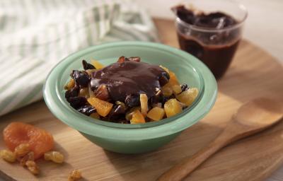 Fotografia em tons de amarelo em uma bancada de madeira de cor marrom. Ao centro, um recipiente verde contendo a salada de frutas. Ao lado, há algumas frutas picadas com uma colher de madeira ao lado. Ao fundo, um pote com a ganache de chocolate.