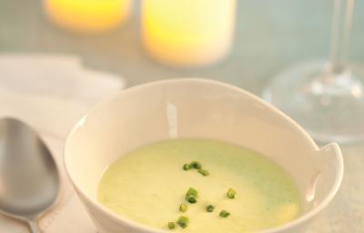 Fotografia em tons de verde em uma mesa de madeira com uma toalha verde, ao centro um potinho branco redondo fundo com o creme de abobrinha com cebolinha decorando. Ao fundo, duas velas acesas e uma taça de vidro com água.