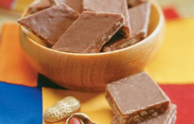 fotografia em tons de marrom, azul e amarelo. Contém um pote redondo marrom com pedaços quadrados de amendoim com chocolate.