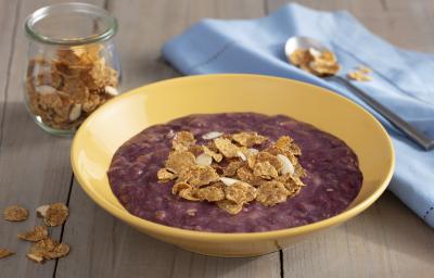 Fotografia em tons de roxo em uma bancada de madeira escura com um pano azul claro ao lado, um prato redondo fundo amarelo com o creme de açaí dentro dele e decorado com cereais. Ao fundo, um potinho de vidro com cereais. Fotografia em tons de roxo em uma bancada de madeira escura com um pano azul claro ao lado, um prato redondo fundo amarelo com o creme de açaí dentro dele e decorado com cereais. Ao fundo, um potinho de vidro com cereais.