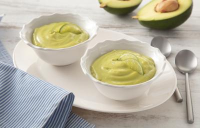 Fotografia em tons de branco e verde de uma bancada, ao centro dois potinhos com creme verde de abacate. Ao fundo, um abacate cortado ao meio. Fotografia em tons de branco e verde de uma bancada, ao centro dois potinhos com creme verde de abacate. Ao fundo, um abacate cortado ao meio.