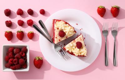 Fotografia vista de cima de um prato raso grande branco de vidro com duas fatias de torta red velvet, com dois garfos juntos ao lado. Ao redor do prato tem mais dois garfos e muitas frutas vermelhas. Fotografia vista de cima de um prato raso grande branco de vidro com duas fatias de torta red velvet, com dois garfos juntos ao lado. Ao redor do prato tem mais dois garfos e muitas frutas vermelhas.