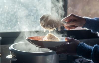 Fotografia de uma mão feminina colocando com uma colher de pau um arroz cremoso ao champanhe em um prato fundo. A imagem está contra a luz do sol, e se caracteriza pelo vapor saindo da panela.