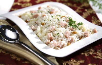 Fotografia em tons de vinho em uma mesa com uma toalha vinho e dourada, um recipiente ao centro branco com o arroz tropical com presunto e salsinha dentro dele. Ao lado, um guardanapo de pano bege com uma colher apoiada nele.
