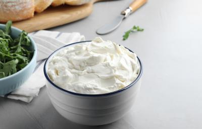 Fotografia em tons de branco com um pote branco ao centro. Dentro do pote existe uma receita de cream cheese caseiro, um creme branco feito com vinagre, leite e creme de leite.