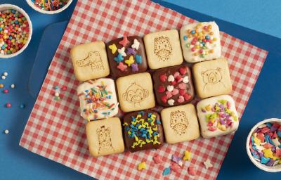 Fotografia em tons de azul e vermelho de uma bancada azul com paninho xadrez vermelho e branco sobre ele biscoitos Passatempo cobertos com chocolate e confeitos coloridos. Ao lado três recipientes brancos redondos com confeitos coloridos. Fotografia em tons de azul e vermelho de uma bancada azul com paninho xadrez vermelho e branco sobre ele biscoitos Passatempo cobertos com chocolate e confeitos coloridos. Ao lado três recipientes brancos redondos com confeitos coloridos.