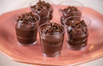 Fotografia em tons de rosa com 5 copinhos de plástico ao centro. Dentro de capa copo existe um brigadeiro de colher de cor marrom escura. Fotografia em tons de rosa com 5 copinhos de plástico ao centro. Dentro de capa copo existe um brigadeiro de colher de cor marrom escura.