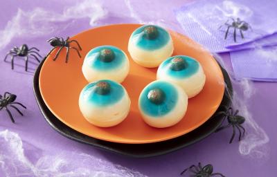 Foto em tons de laranja da receita de olhos de gelatina com Leite moça para o dia das Bruxas servida em uma tábua circular laranja sobre uma mesa roxa com aranhas de decoração