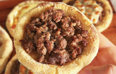 Fotografia mostra uma mão segurando uma esfiha aberta, dourada, com massa macia e levemente crocante nas bordas, recheadas com carne.