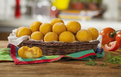 Fotografia em tons de marrom e vermelho em uma bancada de madeira com uma cesta oval de vime e vários bolinhos de abóbora assados com queijo. Ao lado, tomates inteiros e um pedaço de tomate. Fotografia em tons de marrom e vermelho em uma bancada de madeira com uma cesta oval de vime e vários bolinhos de abóbora assados com queijo. Ao lado, tomates inteiros e um pedaço de tomate.