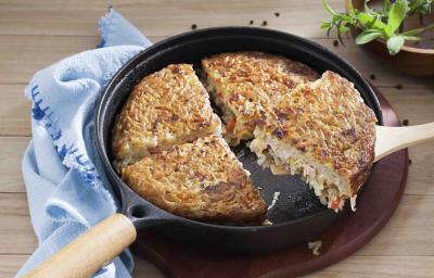 Fotografia em tons de azul em uma bancada de madeira clara com um pano azul, uma tábua de madeira escura, uma frigideira grande preta com a batata rosti recheada com legumes. Fotografia em tons de azul em uma bancada de madeira clara com um pano azul, uma tábua de madeira escura, uma frigideira grande preta com a batata rosti recheada com legumes.