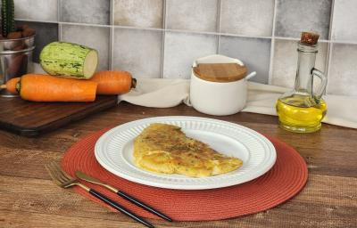 Fotografia em tons de verde e laranja em uma bancada de madeira escura com um apoiador de prato vermelho e um prato branco em cima com a crepioca de legumes. Ao fundo, abobrinha e cenoura cortadas ao meio e vidro de azeite e sal.