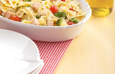 Fotografia em tons de amarelo em uma mesa de madeira com um pano quadriculado em vermelho, um recipiente oval branco com a salada de macarrão. Ao lado, um potinho com azeite.