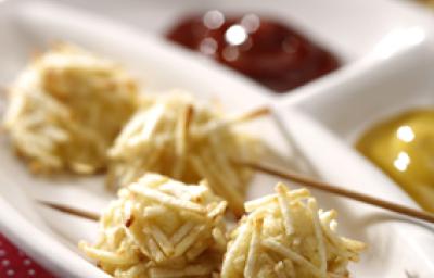 Fotografia em tons de vermelho em uma mesa com uma toalha vermelha com bolinhas brancas, um recipiente branco com o espetinho de cachorro quente empanado com batata palha, e ketchup e mostarda ao lado. Fotografia em tons de vermelho em uma mesa com uma toalha vermelha com bolinhas brancas, um recipiente branco com o espetinho de cachorro quente empanado com batata palha, e ketchup e mostarda ao lado.