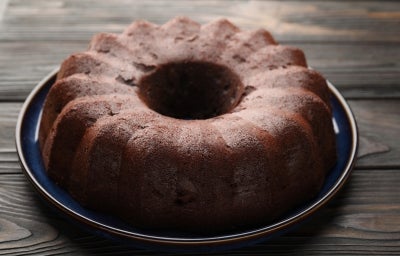 Bolo de chocolate em forma de bundt, com cor marrom escuro, está sobre um prato cerâmico azul. A superfície texturizada sugere umidade, e a apresentação rústica é complementada pela madeira do fundo. Bolo de chocolate em forma de bundt, com cor marrom escuro, está sobre um prato cerâmico azul. A superfície texturizada sugere umidade, e a apresentação rústica é complementada pela madeira do fundo.