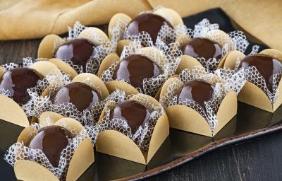 Fotografia em tons de marrom em uma bancada de madeira escura com brigadeiros caramelizados colocados em forminhas beges e ao lado um pano bege.