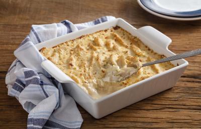 Fotografia em tons de branco e dourado, no centro um refratário com peito de frango gratinado com molho branco e uma colher de servir, ao lado, um guardanapo branco com listras em tons de azul, a borda de um prato, tudo sobre bancada de madeira rústica. Fotografia em tons de branco e dourado, no centro um refratário com peito de frango gratinado com molho branco e uma colher de servir, ao lado, um guardanapo branco com listras em tons de azul, a borda de um prato, tudo sobre bancada de madeira rústica.