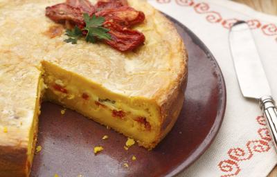 fotografia em tons de marrom e branco de uma bancada marrom vista de cima, contém um pano branco com um prato redondo marrom com a torta faltando uma fatia e por cima tomate seco para decorar. fotografia em tons de marrom e branco de uma bancada marrom vista de cima, contém um pano branco com um prato redondo marrom com a torta faltando uma fatia e por cima tomate seco para decorar.