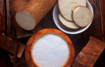 fotografia em tons de marrom e branco de uma bancada marrom vista de cima, contém um recipiente redondo marrom com a farinha dentro e ao lado um recipiente redondo branco com pedaços de mandioca. Em volta contém mandiocas. fotografia em tons de marrom e branco de uma bancada marrom vista de cima, contém um recipiente redondo marrom com a farinha dentro e ao lado um recipiente redondo branco com pedaços de mandioca. Em volta contém mandiocas.