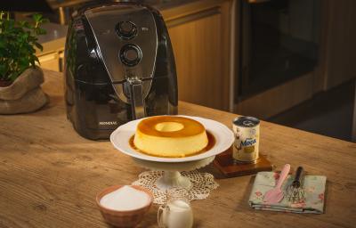 Fotografia em tons de amarelo em uma bancada de madeira, um suporte para bolo branco e o pudim e Leite Moça em cima dele. Ao fundo, uma air fryer Mondial, uma lata de leite Moça, um paninho azul florido com uma espátula rosa e um fouet em cima.