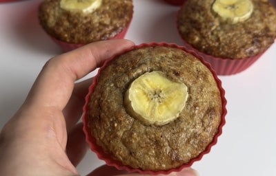 Fotografia de uma mão segurando um muffin de banana com aviea em uma forminha de silicone. No fundo tem mais muffins sobre uma mesa branca.