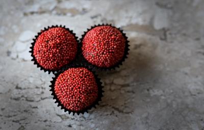 Fotografia em tons de vermelho, em uma bancada de madeira de cor cinza. Ao centro, 3 formilhas de brigadeiro contendo os brigadeiros. Fotografia em tons de vermelho, em uma bancada de madeira de cor cinza. Ao centro, 3 formilhas de brigadeiro contendo os brigadeiros.