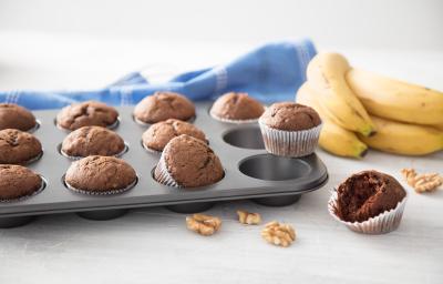 Fotografia em tons de cinza, marrom e amarelo, forma com cupcakes de chocolate em bancada cinza, nozes espalhadas ao redor, ao lado um cacho de banana e um cupcake aberto e ao fundo um guardanapo azul com listras brancas.