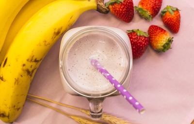 Fotografia em tons de rosa, de uma bancada rosa vista de cima, ao centro um copo com uma bebida e um canudo, em volta bananas e morangos. Fotografia em tons de rosa, de uma bancada rosa vista de cima, ao centro um copo com uma bebida e um canudo, em volta bananas e morangos.