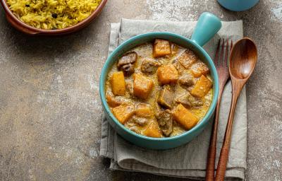 Fotografia em tons de marrom em uma bancada de madeira de cor cinza. Ao centro, um pano cinza contendo uma panela azul em cima com o cozido dentro. Ao lado, uma colher e um garfo de madeira e ao fundo um recipiente contendo arroz amarelo. Fotografia em tons de marrom em uma bancada de madeira de cor cinza. Ao centro, um pano cinza contendo uma panela azul em cima com o cozido dentro. Ao lado, uma colher e um garfo de madeira e ao fundo um recipiente contendo arroz amarelo.
