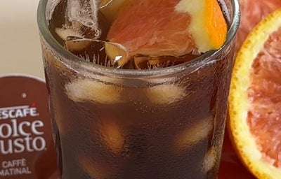 Fotografia de um copo de vidro com uma bebida gelada de nescafé e suco de laranja, dentro do copo também tem cubos de gelo e um pedacinho de laranja. Ao redor do copo tem uma cápsula de nescafé dolce gusto usada na receita e laranjas abertas. Fotografia de um copo de vidro com uma bebida gelada de nescafé e suco de laranja, dentro do copo também tem cubos de gelo e um pedacinho de laranja. Ao redor do copo tem uma cápsula de nescafé dolce gusto usada na receita e laranjas abertas.