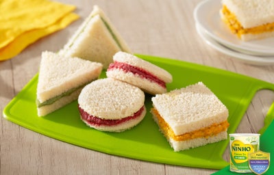 Sanduiches variados de pão branco com recheios coloridos, em formatos redondo, triangular e quadrado.