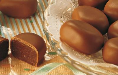 Fotografia em tons de marrom, em uma bancada de madeira com um suporte para bolo de vidro e vários bombons de chocolate em cima dele. Fotografia em tons de marrom, em uma bancada de madeira com um suporte para bolo de vidro e vários bombons de chocolate em cima dele.