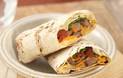 Fotografia em tons de marrom em uma bancada de madeira com uma tábua de madeira clara, um prato redondo fundo com dois wraps de carne com legumes. Fotografia em tons de marrom em uma bancada de madeira com uma tábua de madeira clara, um prato redondo fundo com dois wraps de carne com legumes.