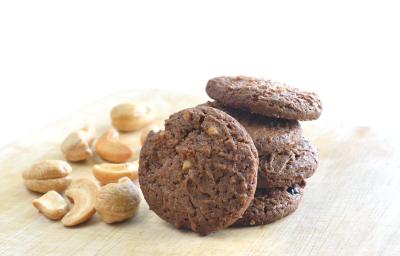 fotografia em tons de branco, marrom e bege de uma bancada bege vista de frente, ao centro biscoitos de chocolate e ao lado castanhas de caju