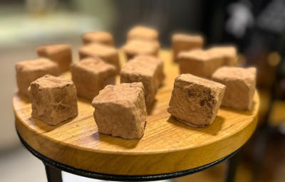 Fotografia em tons de marrom com uma bancada de madeira ao centro. Em cima da bancada existem vários quadrados de chocolate cobertos com chocolate em pó Fotografia em tons de marrom com uma bancada de madeira ao centro. Em cima da bancada existem vários quadrados de chocolate cobertos com chocolate em pó